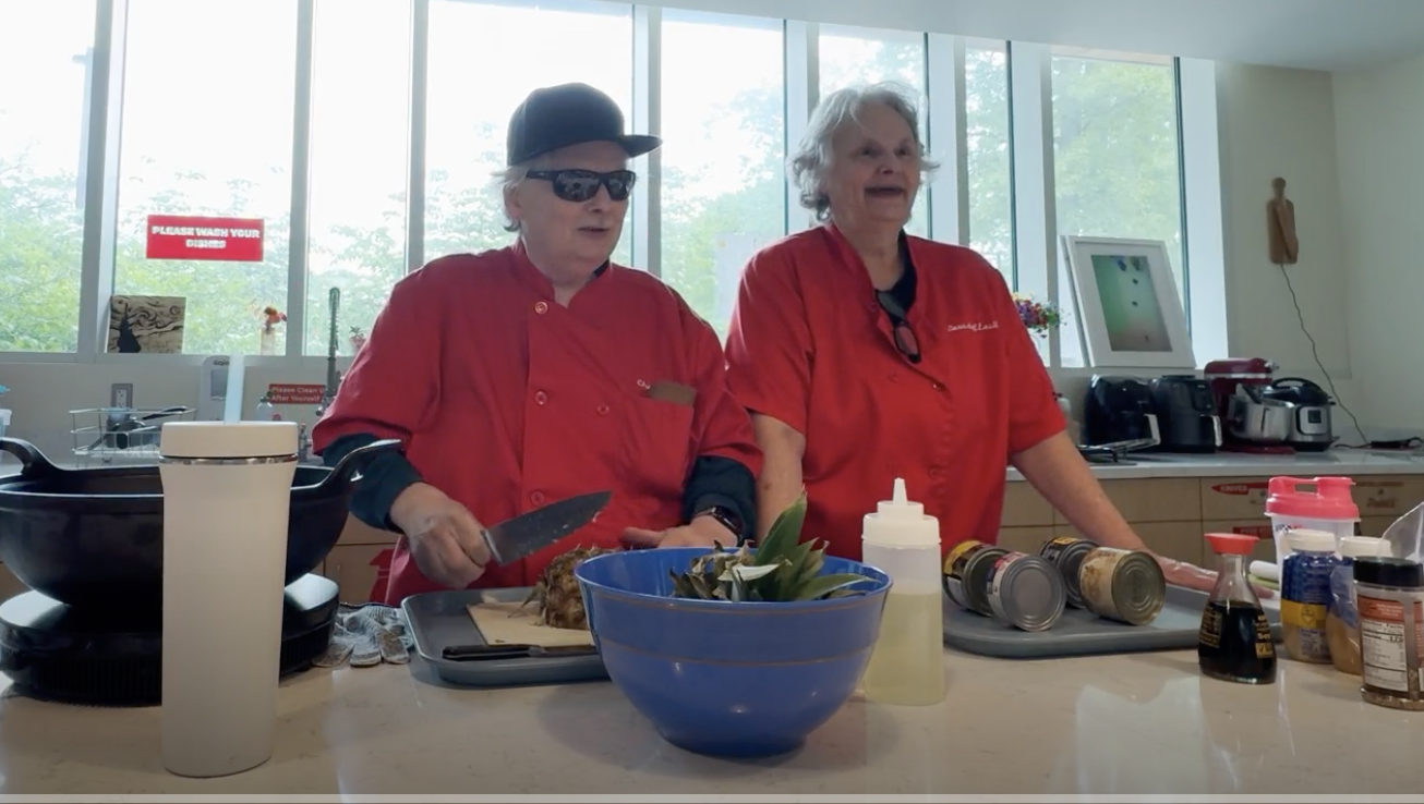 Brian Charlson and Lesli Charlson cooking. Brian is holding a large knife and about to chop a pineapple